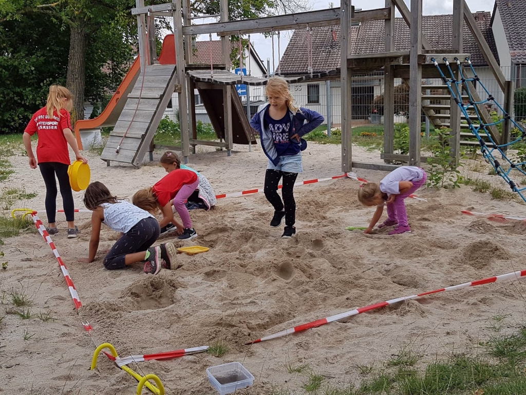 Ferienerlebnistag beim TSV Wiernsheim 10 Kinder im Sandspielbereich mit Schaufeln und roten Markierungsstangen, während sie Formen in den Sand graben und befüllen.
