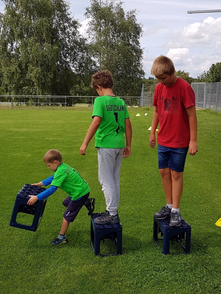 Ferienerlebnistag beim TSV Wiernsheim 11 Drei Kinder stehen auf Fußballtrainingshockern im Rasen, schauen nach unten und üben Bewegungsabläufe an einem Outdoor-Sportplatz.