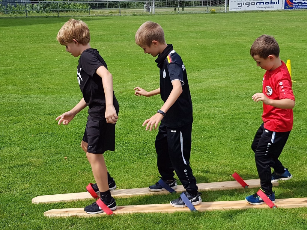 Ferienerlebnistag beim TSV Wiernsheim 12 Drei Kinder auf dem Kunstrasen, üben Schrittfolgen auf mit Markierbändern begrenzten Holzbahnen für Koordination und Gleichgewicht.