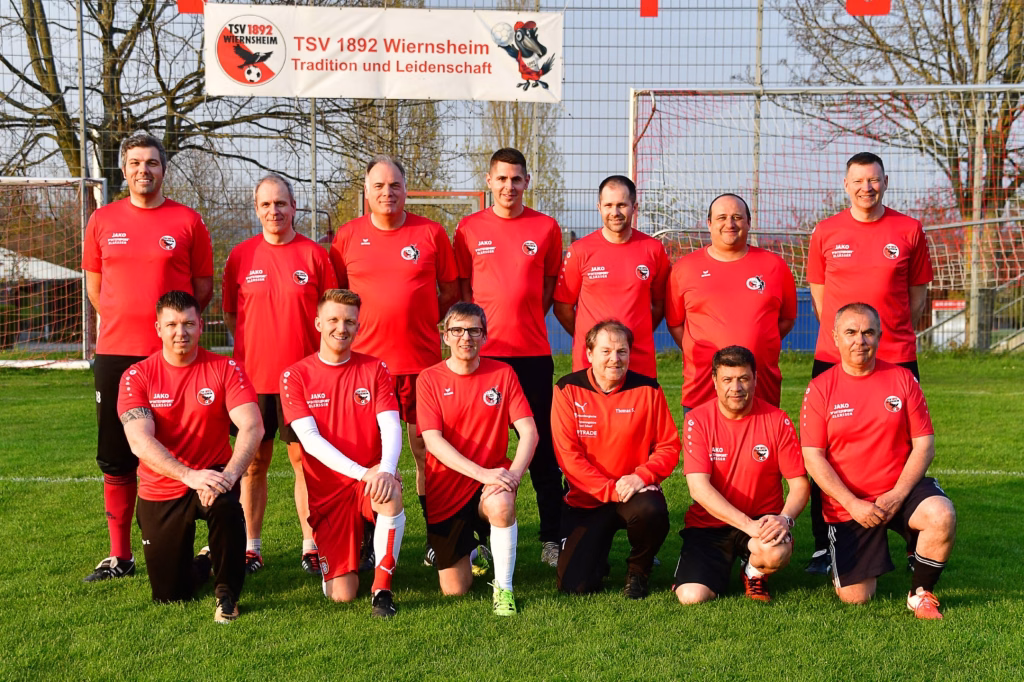 Mannschaftsfoto auf dem Fußballplatz: mehrere Personen in roten Trikots, stehend und kniend vor Tor und Fangnetz, mit Mannschaftsbanner im Hintergrund.