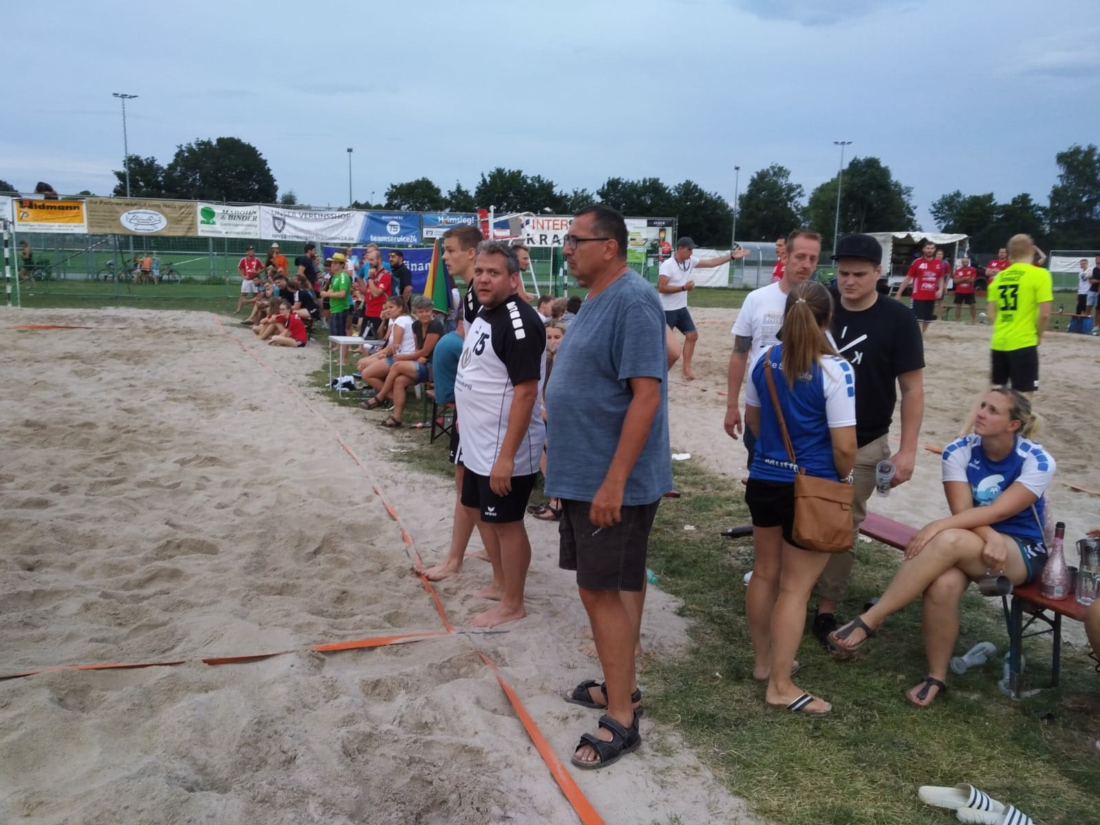Zuschauer bei einem Beachsport-Turnier auf dem Sandfeld; Helfer und Teilnehmer stehen am Rand einer umrandeten Spielfläche.