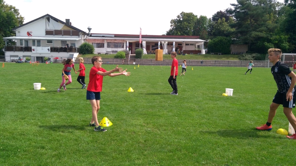 Ferienerlebnistag beim TSV Wiernsheim 13 Kinder mit roten Sportshirts üben auf einem Rasenparcours mit Trikots, Kegeln und Markierungsbechern Bewegungs- und Zielübungen.