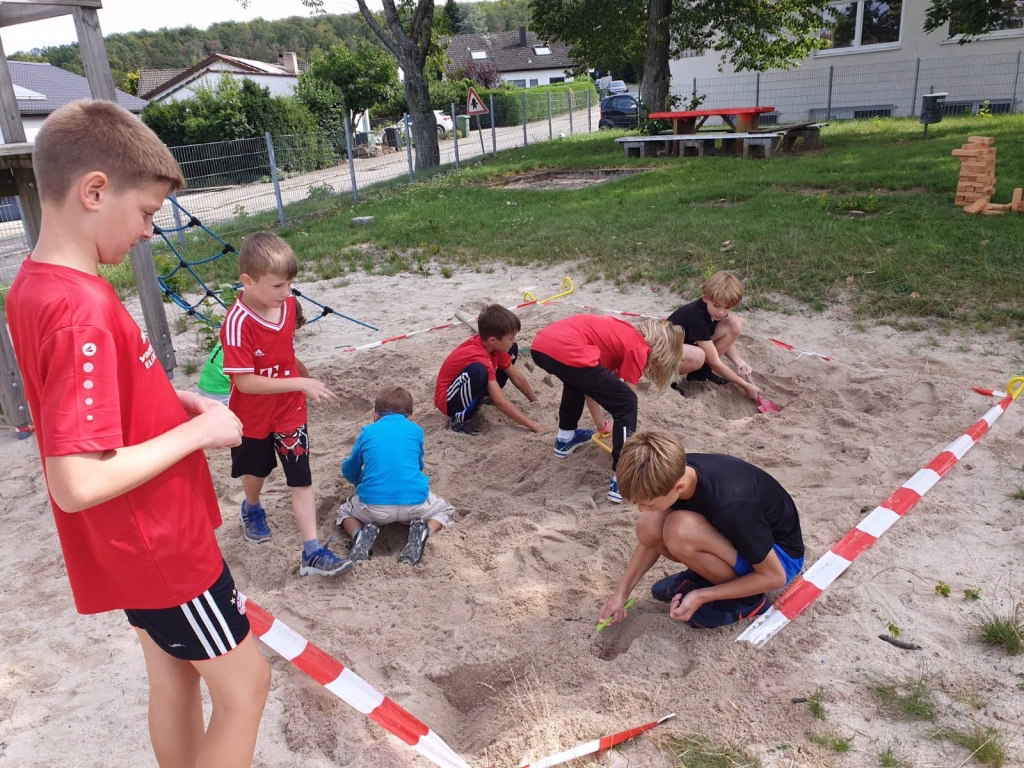 Ferienerlebnistag beim TSV Wiernsheim 15 Kinder spielen im Sandkasten und buddeln mit Händen und Werkzeugen entlang roter Markierungsbänder, im Garten vor einem Zaun.