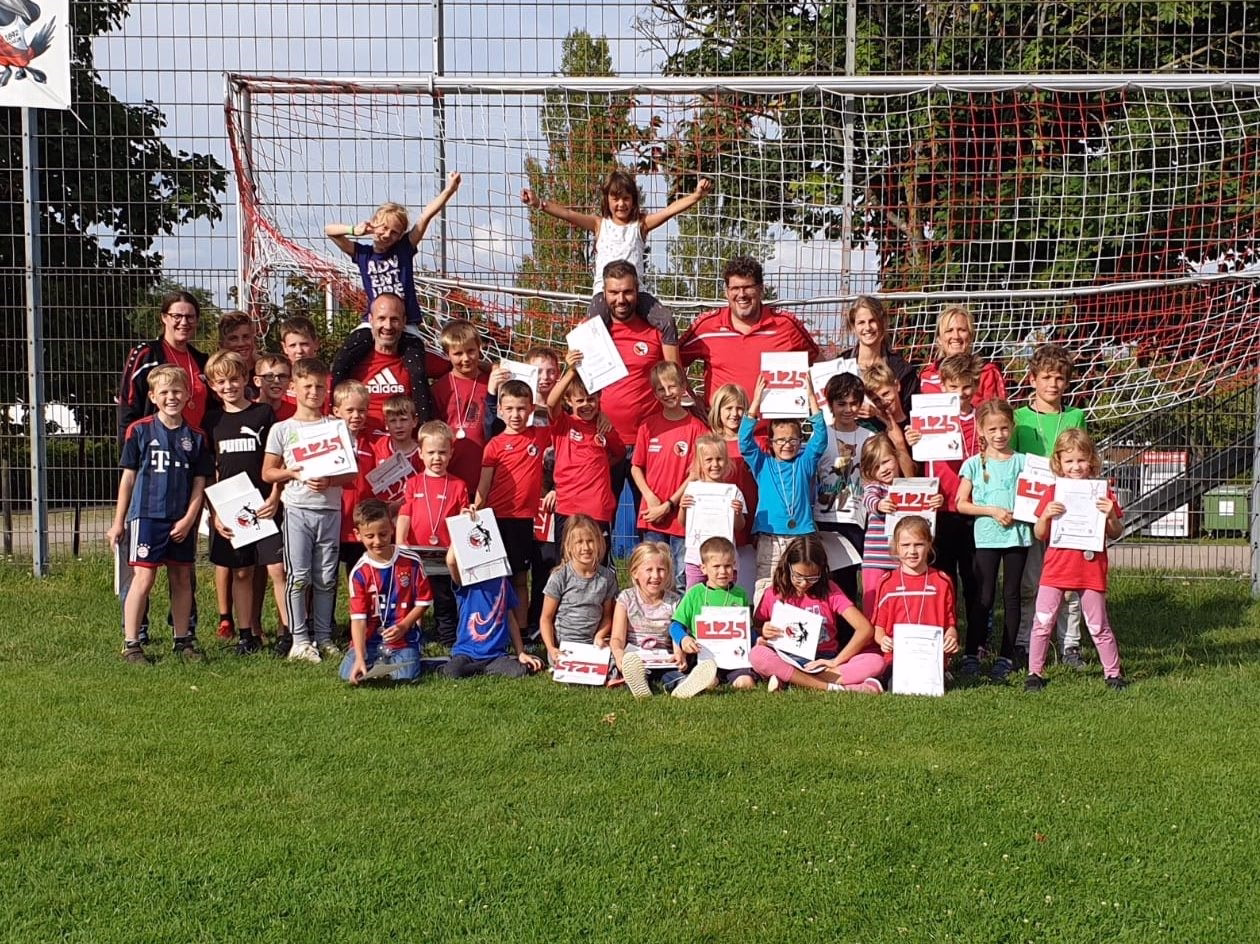 Gruppenfoto von Kindern und Trainern vor einem Fußballtor auf dem Rasen; alle halten Urkunden zur Sieger- oder Abschlussfeier.