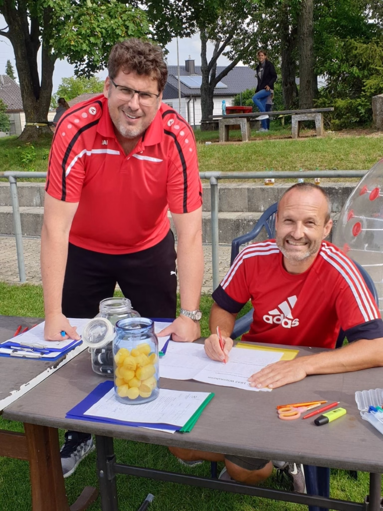 Ferienerlebnistag beim TSV Wiernsheim 16 Zwei Männer am Gartentisch beim Unterschreiben von Formularen, um eine Veranstaltung zu dokumentieren; Stifte, Gläser und Papier liegen bereit.