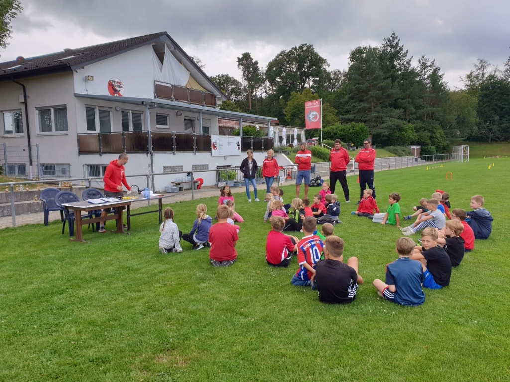 Ferienerlebnistag beim TSV Wiernsheim 3 Kinder sitzen im Kreis auf Rasen vor einem Gebäude; Erwachsene in Sportkleidung geben Erklärungen bei einer Veranstaltung im Freien.