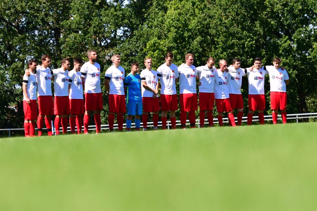 Fußballmannschaft steht im Halbkreis auf dem Trainingsfeld vor grünem Hintergrund und posiert im Freien.