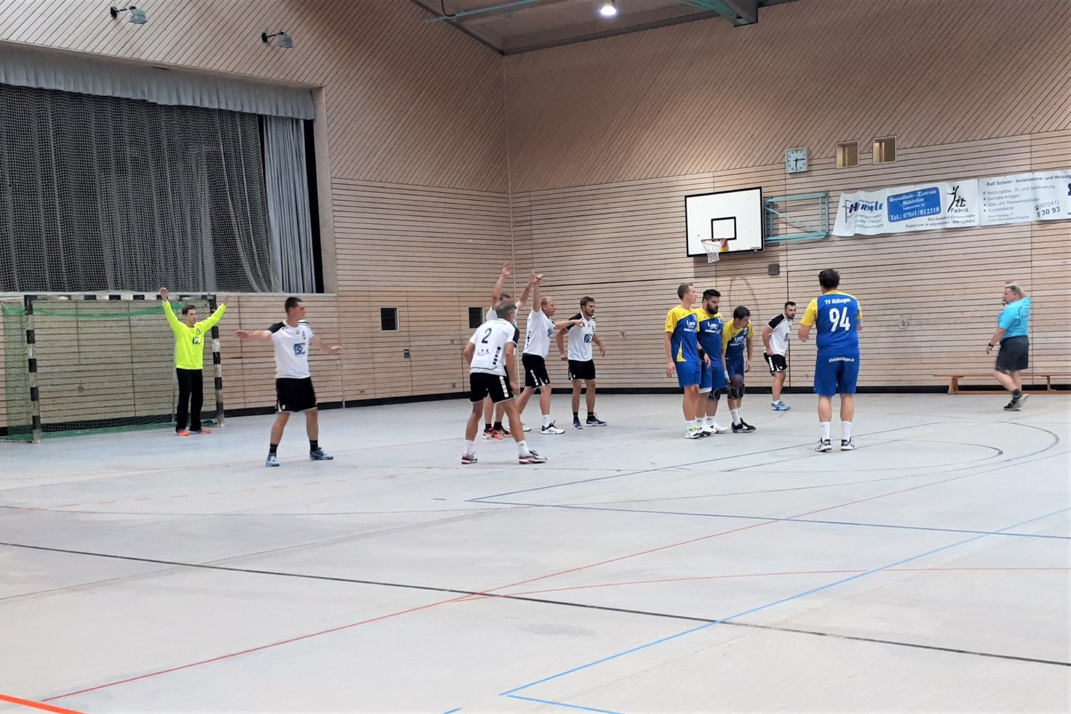 Handballspiel in einer Turnhalle: Spieler stellen sich hintereinander, werfen in Richtung Tor, Schiedsrichter beobachtet von links, Hallenwand mit Korb.