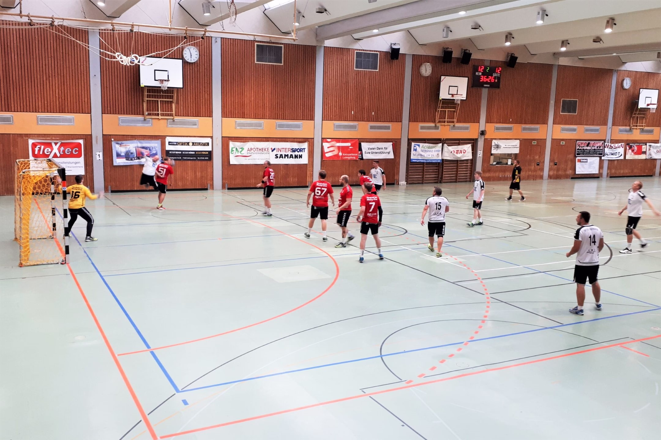 Handballspiel in einer Sporthalle: mehrere Spieler stehen am Kreis, ein Torwart im Kasten, leere Mannschaftsbereiche vor beschrifteten Werbewänden.