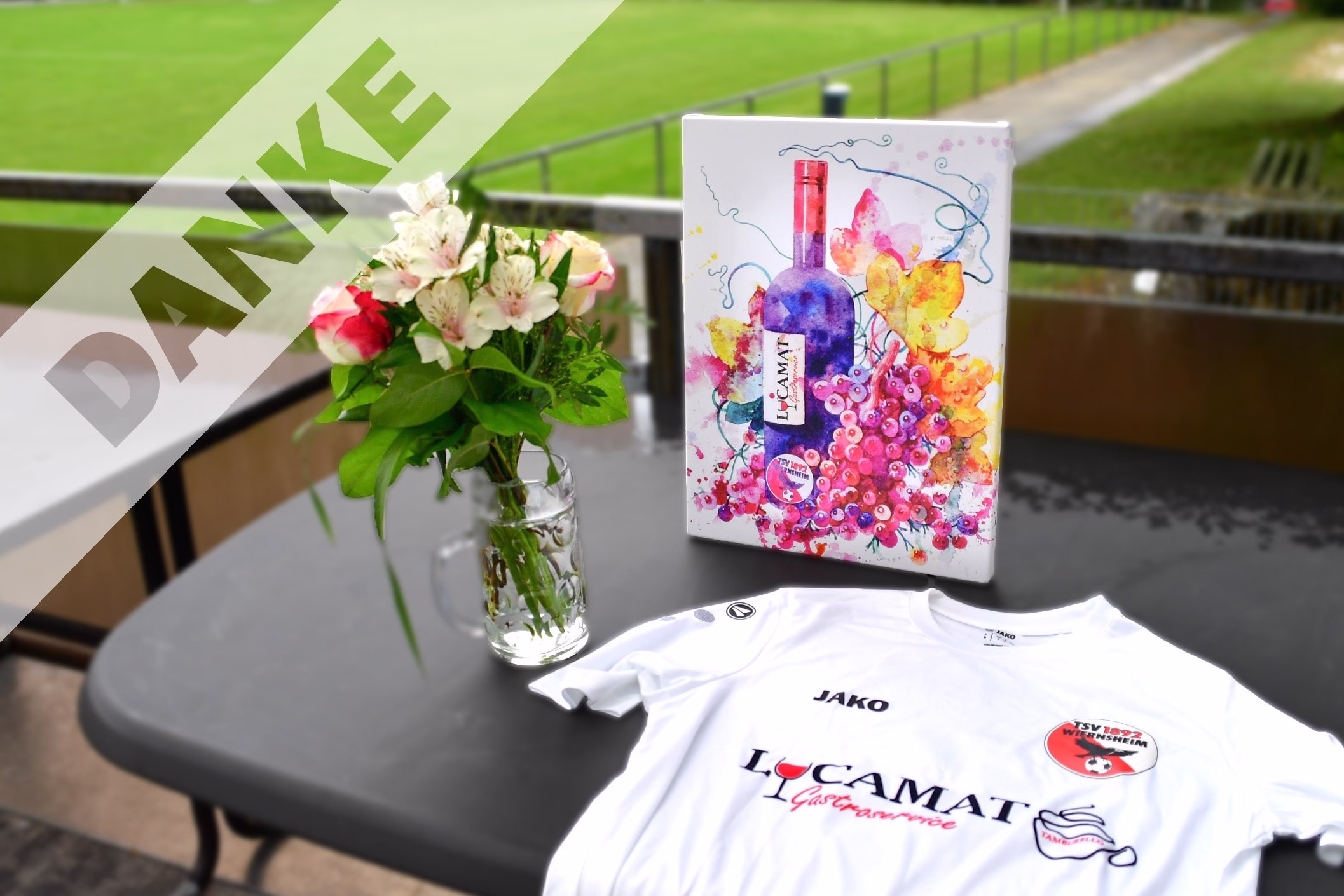 Tischdekoration mit Blumenstrauß in Vase, Präsentationsbox und T-Shirt auf einer Sitzbank, im Hintergrund Rasen und Zaun.