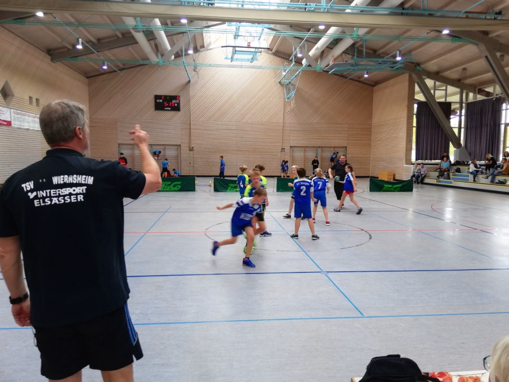 Heimspieltag der männl. E-Jugend 4 Handballspiel in einer Sporthalle: Trainer verfolgt das Geschehen, Kinder in Trikots ringen um den Ball auf dem Spielfeld.