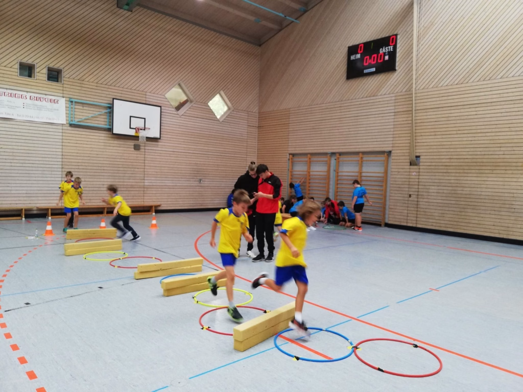 Heimspieltag der männl. E-Jugend 6 Sporthalle mit Kindern beim Parcours-Laufen über Bodenelemente; Kreise und Hütchen markieren Laufwege, Schiedsrichter im Hintergrund.
