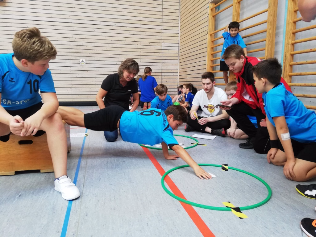 Heimspieltag der männl. E-Jugend 3 Kinder knien in einer Sporthalle auf einem Hallenboden mit farbigen Markierungen und hula-hoop-Reifen und üben eine gemeinsame Übung.