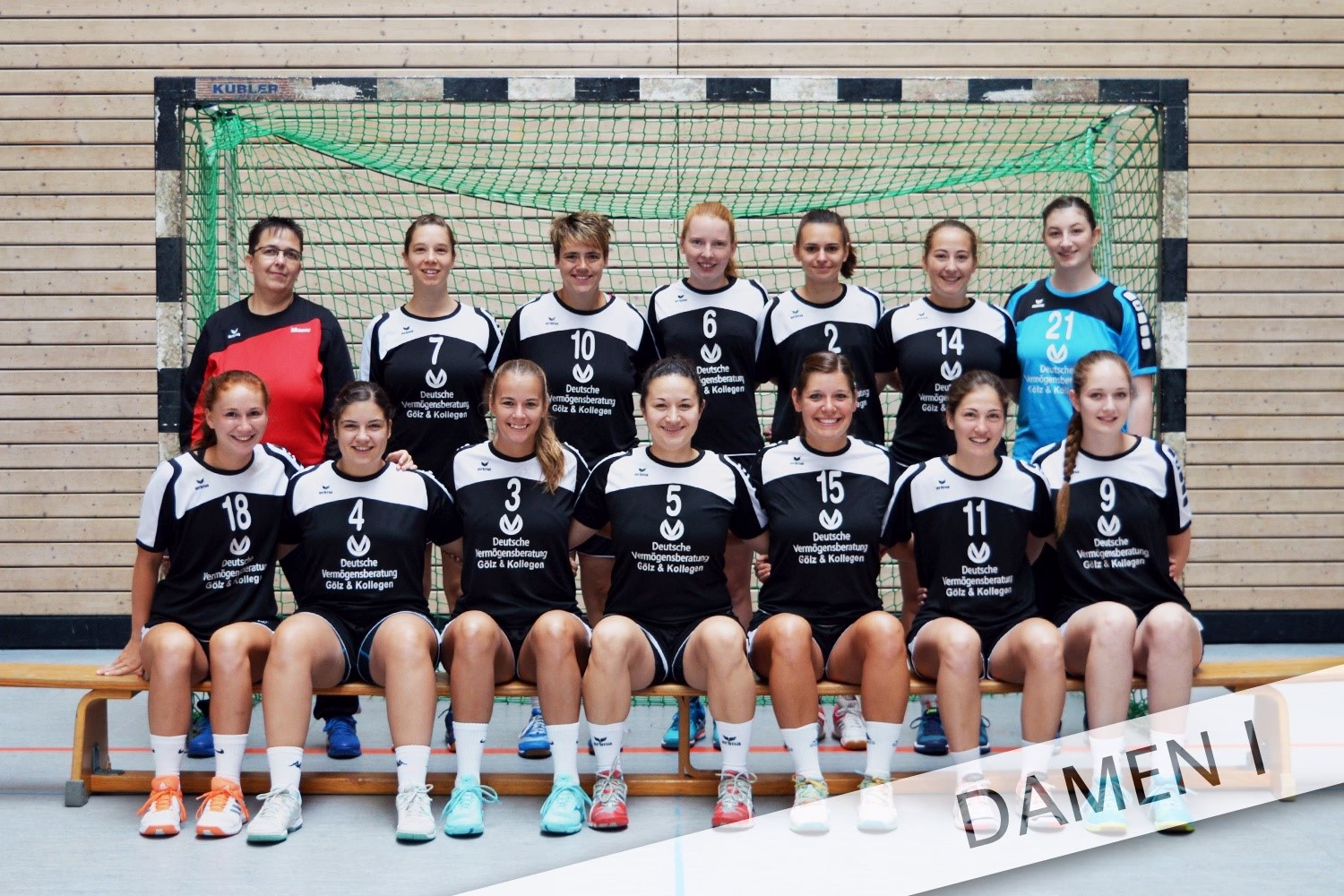 Handball-Team mit mehreren Spielerinnen posiert vor einem Tor; Sporttrikots und Trikotnummern, Teamfoto in Hallenumgebung.