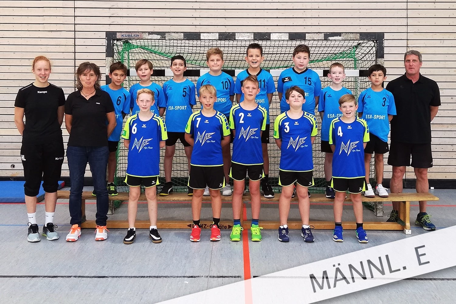 Mannschaftsfoto in Sporthalle: Kinder im Trikot stehen mit Trainer vor einem Tor, nummerierte Vordergrundspieler für Team-Darstellung.