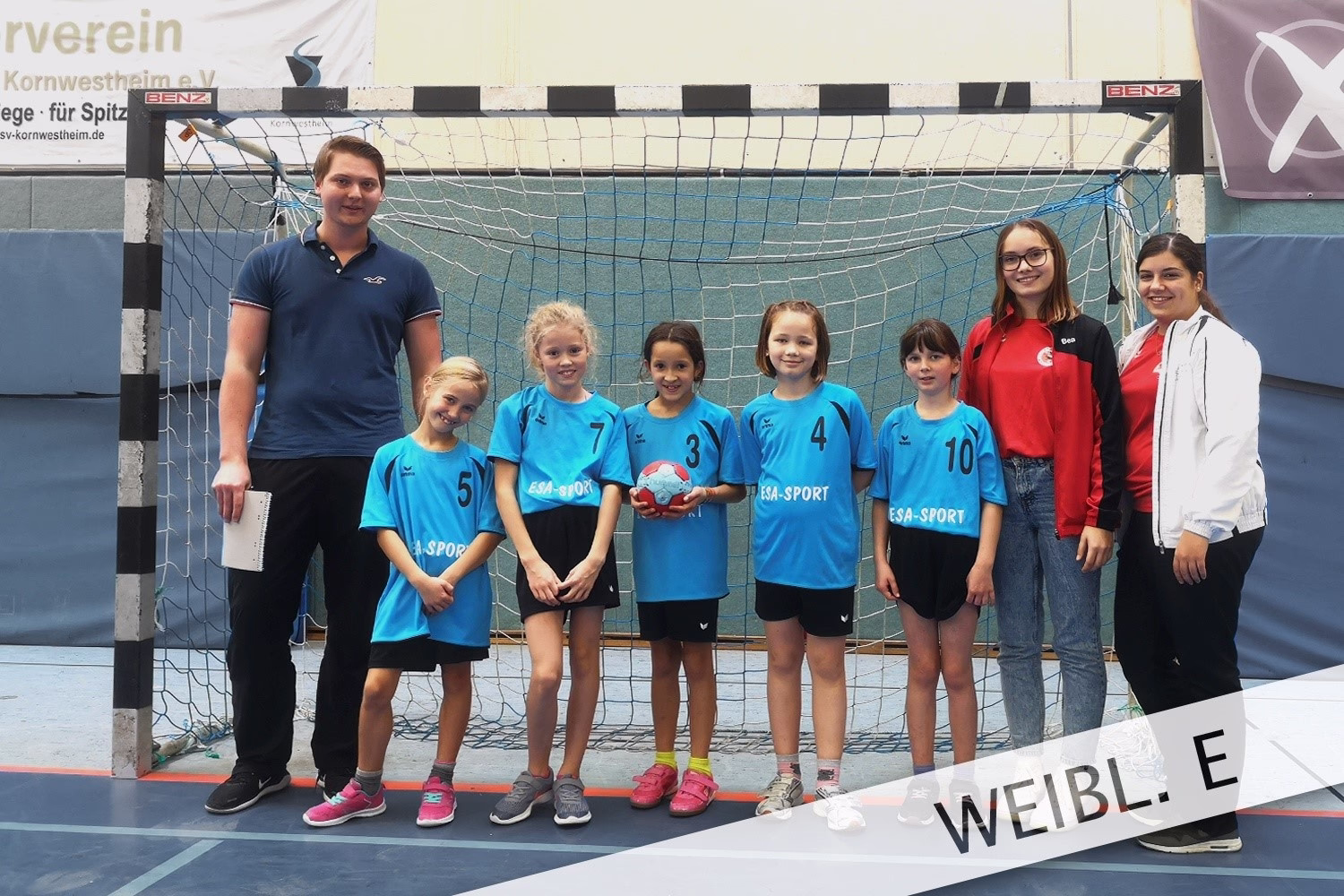 Sportteam vor einem Handballtor: Trainerin und mehrere Mädchen in Trikots, posieren für Mannschaftsfoto.