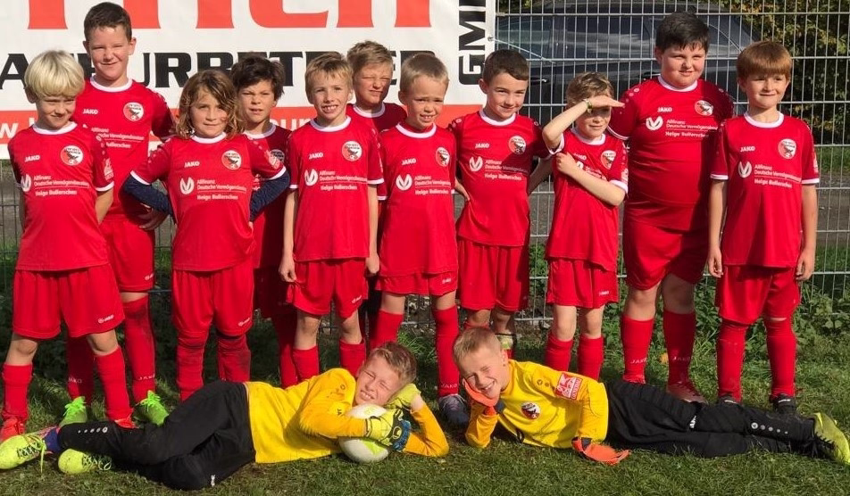 Kinderfußballteam in roten Trikots posiert vor Zaun; zwei Kinder liegen lachend im Vordergrund, andere stehen im Hintergrund.