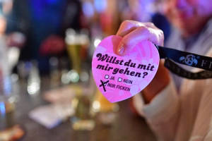 Hand hält ein rosa Herz mit Aufdruck „Willst du mit mir gehen?“, Antwortfelder „Ja“ und „Nein“ sowie „Nur ficken“ an Konfetti-Partybereich.