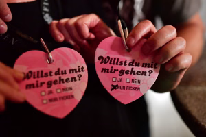 Hände schreiben auf rosa Herz-Karten: „Willst du mit mir gehen?“ mit Antwortkästchen „Ja“/„Nein“ und Hinweis „Nur ficken“.