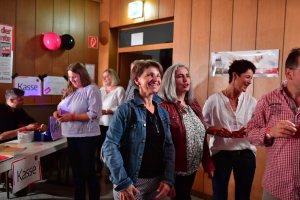 Personengruppe bei Feier im Innenraum, im Vordergrund Lächeln und Gespräche, im Hintergrund Kasse und Ballons als Anlass-Dekoration.