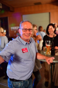 Lächelnder Mann mit Brille und Hemd am Stehtisch auf einer Feier, Hand auf der Tischkante, Gäste im Hintergrund.