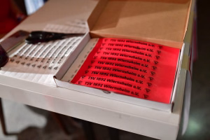 Geöffnete Kartonbox mit rotem Aufdruck; wiederholt gedruckte Vereinsangaben und Emblemzeilen als Etikett für eine Lieferung.