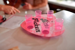 Rosafarbene Gläser auf einer Platte mit Zahlendruck, im Vordergrund; Personen bereiten das Servieren eines Drinks vor.