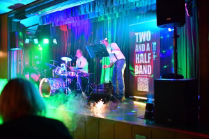 Live-Band auf einer Bühne mit farbigen Lichtern, Sänger am Mikrofon, Schlagzeuger im Hintergrund, Textbanner und QR-Code im Bühnenbild.