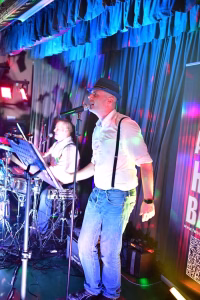 Sänger mit Hut vor Mikrofon auf einer Bühne; Bühnenlicht und Gitarristin hinter Notenständer, um ein Livekonzert zu zeigen.
