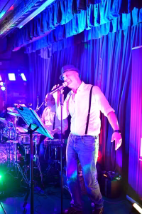 Sänger auf einer Bühne mit Mikrofon, beleuchtet von farbigen Scheinwerfern; Hintergrundvorhänge und Schlagzeug unterstützen das Live-Konzert-Setting.