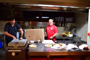 Zwei Personen in einer Küchenzeile beim Zubereiten und Portionieren von Essen an einem Servierwagen mit Schüsseln und Schalen.