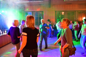 Personen feiern in einem Saal; Lichtfarben beleuchten eine Bühne, im Vordergrund stehen Zuschauer, im Hintergrund spricht/dirigiert eine Person.