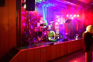 Musiker auf einer Bühne mit Schlagzeug und Keyboard, beleuchtet von farbigen Lichtstrahlen, im Hintergrund ein farbiges Banner.