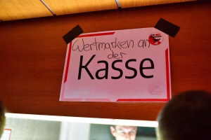 Schild mit rotem Rand und Schrift „Werktwagen an der Kasse“, aufgehängt an einer Holzdecke, markiert den Ort der Kasse.