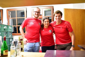 Drei Personen in roten Sportshirts posieren im Innenraum vor einem Tresen, lächeln in die Kamera; Trophäen und Flaschen im Vordergrund.