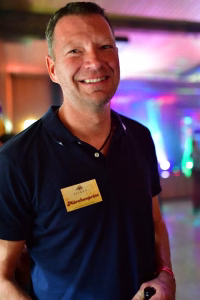 Lächelnder Mann im schwarzen Poloshirt mit Namensschild bei einer festlichen Veranstaltung vor buntem Licht im Hintergrund.