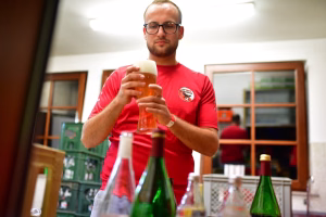 Mann im roten T‑Shirt hält ein Glas mit bernsteinfarbenem Getränk und stößt gemeinsam mit Freunden an; Hintergrund mit Flaschen beim Buffet.