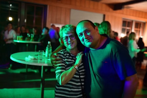 Zwei Personen posieren eng beieinander und lächeln vor einer Party mit grüner Beleuchtung; im Hintergrund steht ein Tisch mit Getränken.