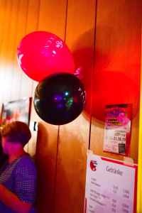 Rote und schwarze Luftballons an einer Wand, im Hintergrund ein Plakat und eine sitzende Person; Deko für eine Feier.