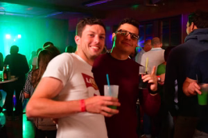 Zwei Gäste feiern in einer beleuchteten Partyumgebung, stoßen mit Getränken an und lächeln in die Kamera.