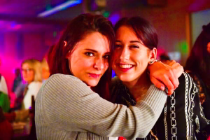 Zwei junge Frauen posieren eng umarmt vor bunten Partylichtern; eine lächelt in die Kamera, die andere blickt ruhig.