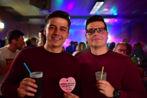 Zwei Männer in rotem Pullover halten Getränke mit Strohhalm und eine rosa Herzkarte; im Hintergrund eine festlich beleuchtete Feiergesellschaft.