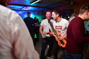 Festveranstaltung mit drei lachenden Männern in Party-Umgebung, neonfarbene Beleuchtung und Getränkepick im Vordergrund.