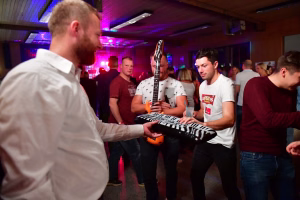 Mann reicht einem Musiker ein großes, bunt bedrucktes Tablett mit Flaschen; Partyraum mit farbigen Lichtern, Gäste schauen dabei zu.