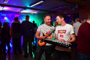 Zwei Männer lachen in einer Party-Lounge; einer hält eine orangefarbene Gitarre, der andere ein Glas, bunte Bühnenbeleuchtung im Hintergrund.
