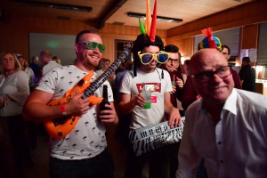 Feiernde Menschen mit Partybrillen und Luftschlangen, eine Person hält eine Gitarre, im Vordergrund ein breites Grinsen beim Anstoßen.