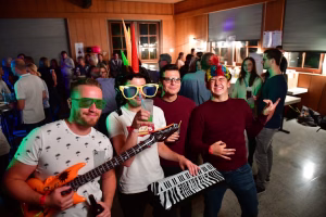 Feiernde Personen im Innenraum mit Partybrillen, buntem Kopfschmuck und Musikinstrumenten; Stimmung einer festlichen Veranstaltung mit Lichtreflexen.