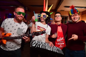 Vier lachende Feiernde mit Partybrillen und Kostümhaaren, einer spielt Gitarre, einer hält bunte Trinkhalme und Gläser in einer festlichen Location.