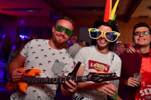 Zwei Feiern­de mit Partybrillen und Schnurrbart-Hut halten eine Gitarre und trinken; ausgelassene Stimmung auf einer Indoor-Party.