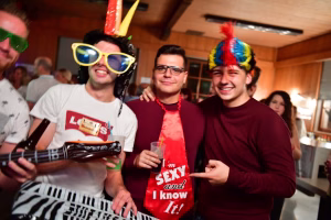 Drei lachende Männer auf einer Party, mit Partyhüten und Getränken; einer hält einen Konfettiknüller, alle feiern gemeinsam.
